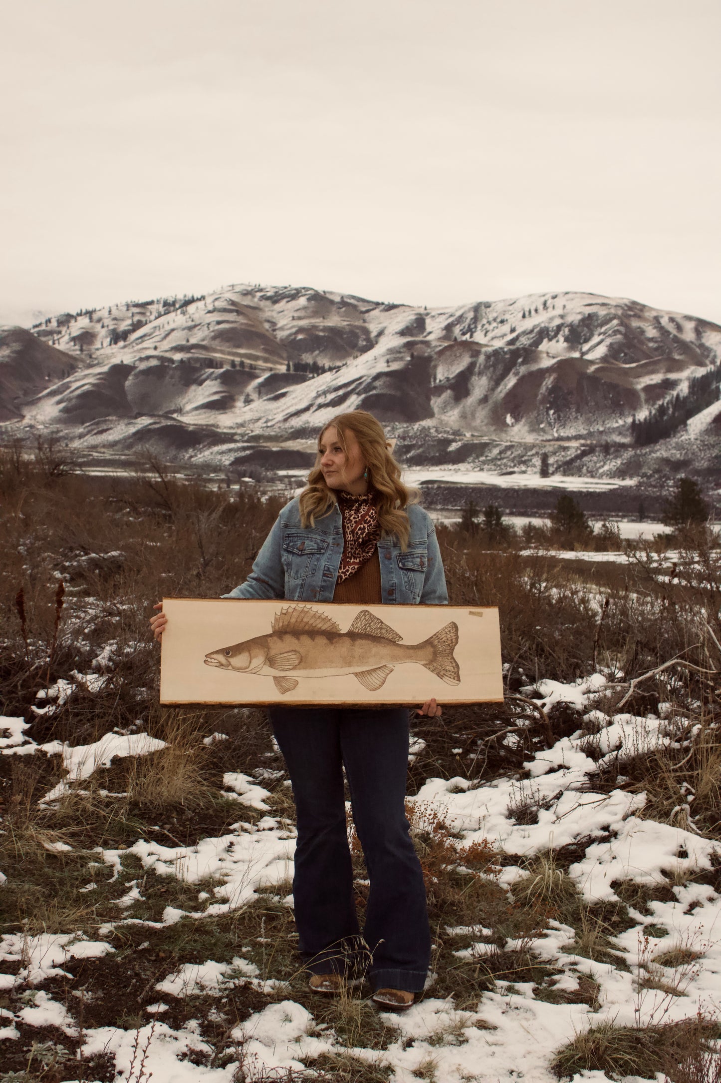 Person holding a painting of a fish in a snowy landscape with mountains.