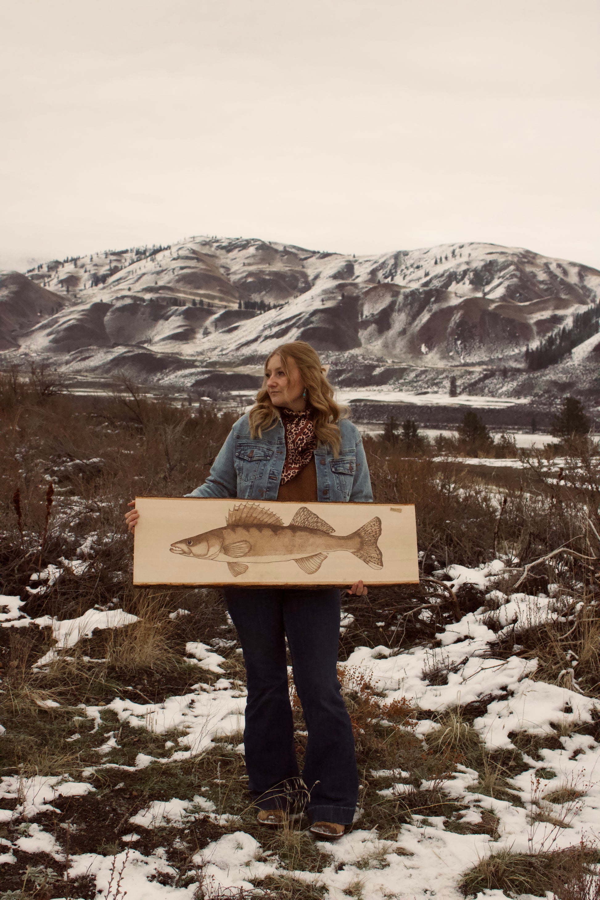 Person holding a painting of a fish in a snowy landscape with mountains.