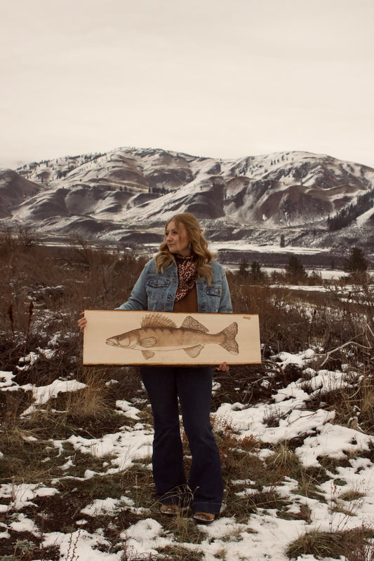Person holding a painting of a fish in a snowy landscape with mountains.